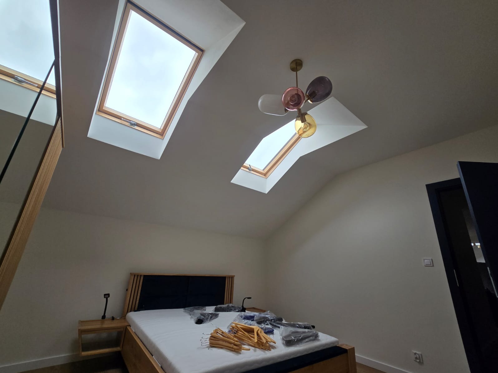 Sypialnia na poddaszu z drewnianym łóżkiem, trzema oknami dachowymi i nowoczesną lampą z kolorowymi kloszami, w trakcie urządzania, z folią ochronną na materacu i stertą drewnianych wieszaków.
