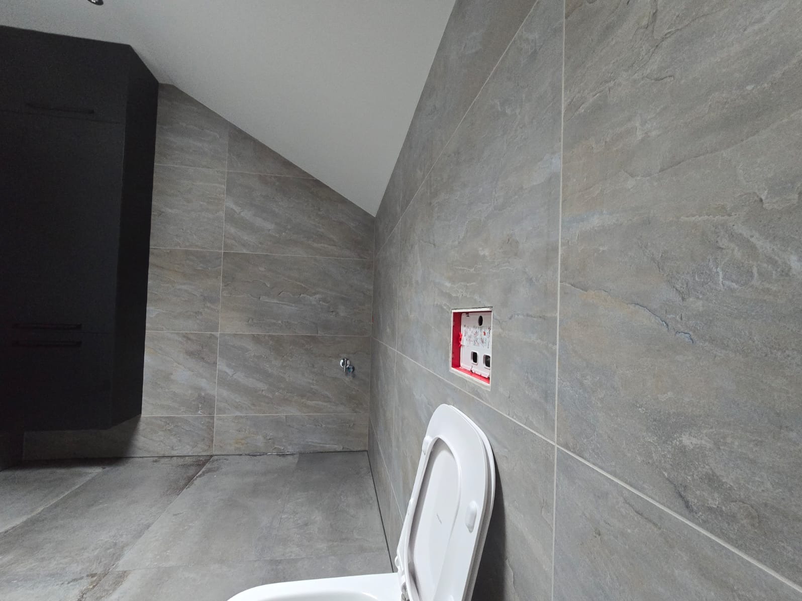 Szara łazienka w trakcie wykańczania: płytki imitujące kamień na ścianach i podłodze, widoczna instalacja podtynkowa, częściowo zamontowana toaleta i czarna szafka.