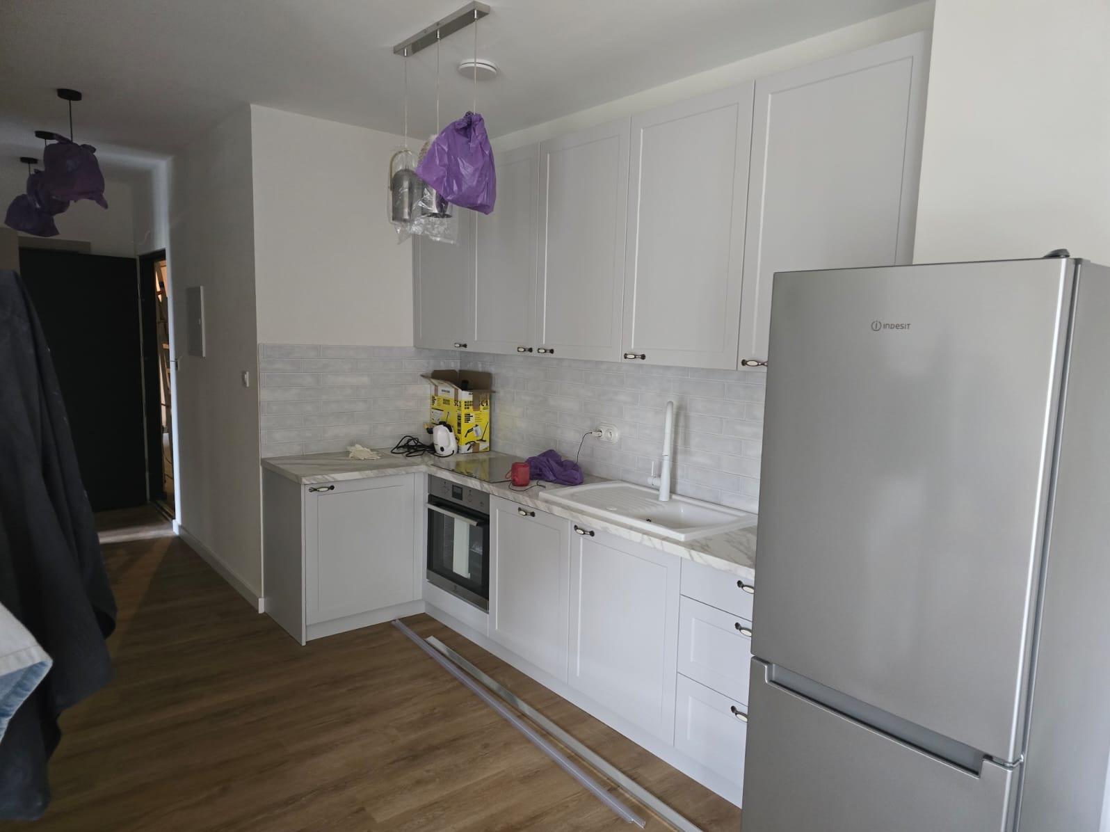 Jasna kuchnia z białymi szafkami, szarymi płytkami imitującymi cegłę i srebrną lodówką, z widocznymi elementami montażowymi na podłodze i lampami wiszącymi w folii ochronnej.