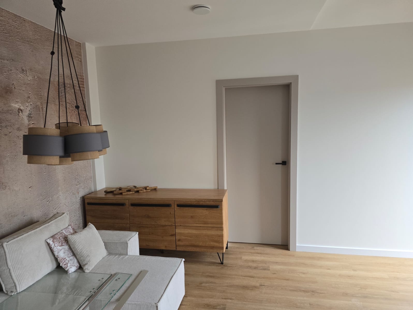 Nowoczesny salon z drewnianą komodą, szarym narożnikiem, designerską lampą wiszącą i beżowymi ścianami oraz podłogą.