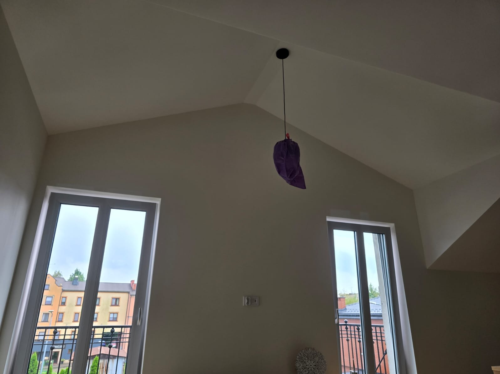 Pomalowane na jasno ściany w pomieszczeniu ze skośnym sufitem, z dwoma balkonowymi oknami po bokach i wiszącą na kablu lampą zabezpieczoną fioletowym workiem.