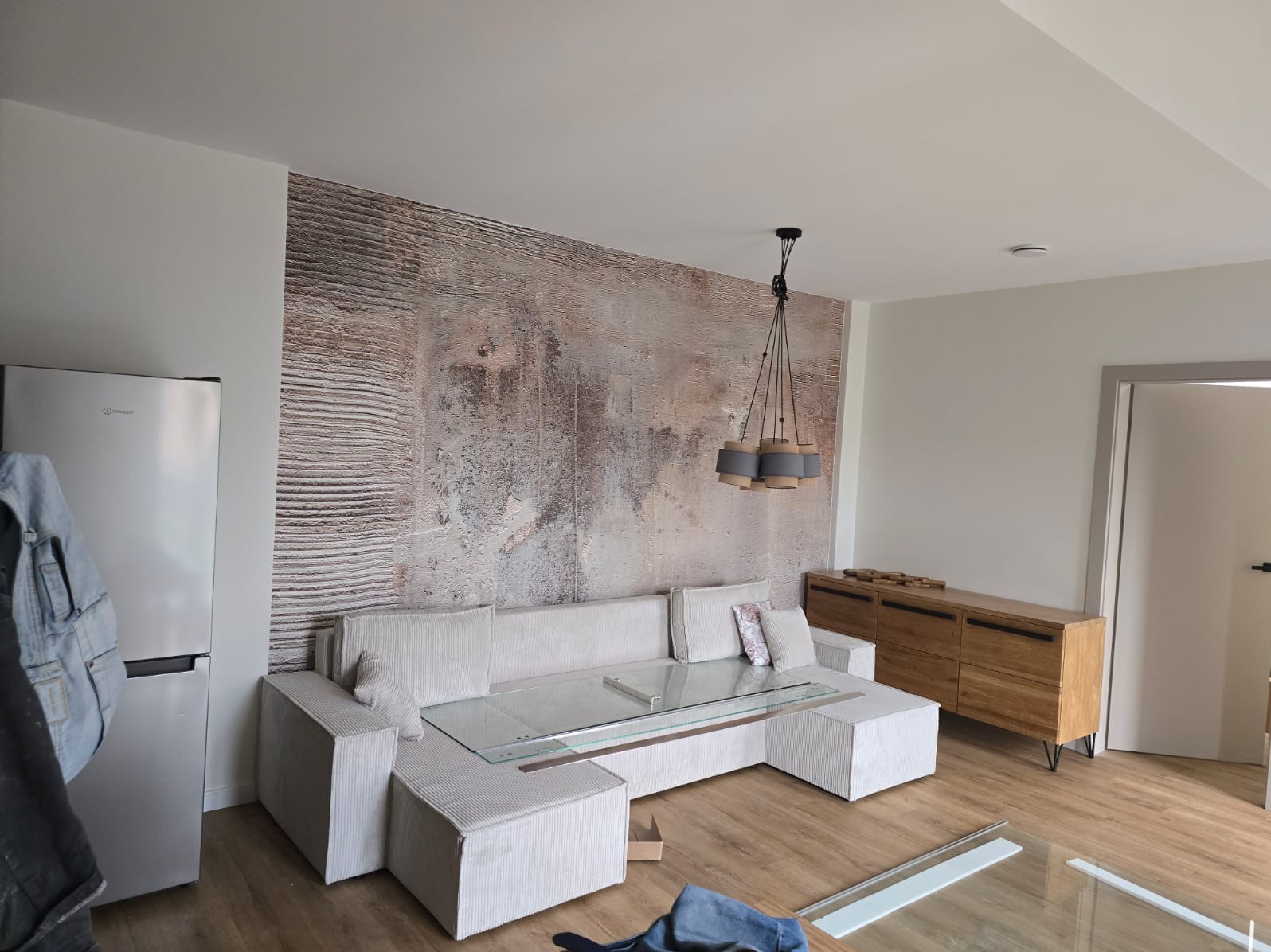 Salon z narożną sofą z jasnej tkaniny, szklanym stolikiem kawowym i ścianą z dekoracyjną tapetą o fakturze drewna, oświetlony wiszącą lampą z szarymi abażurami.