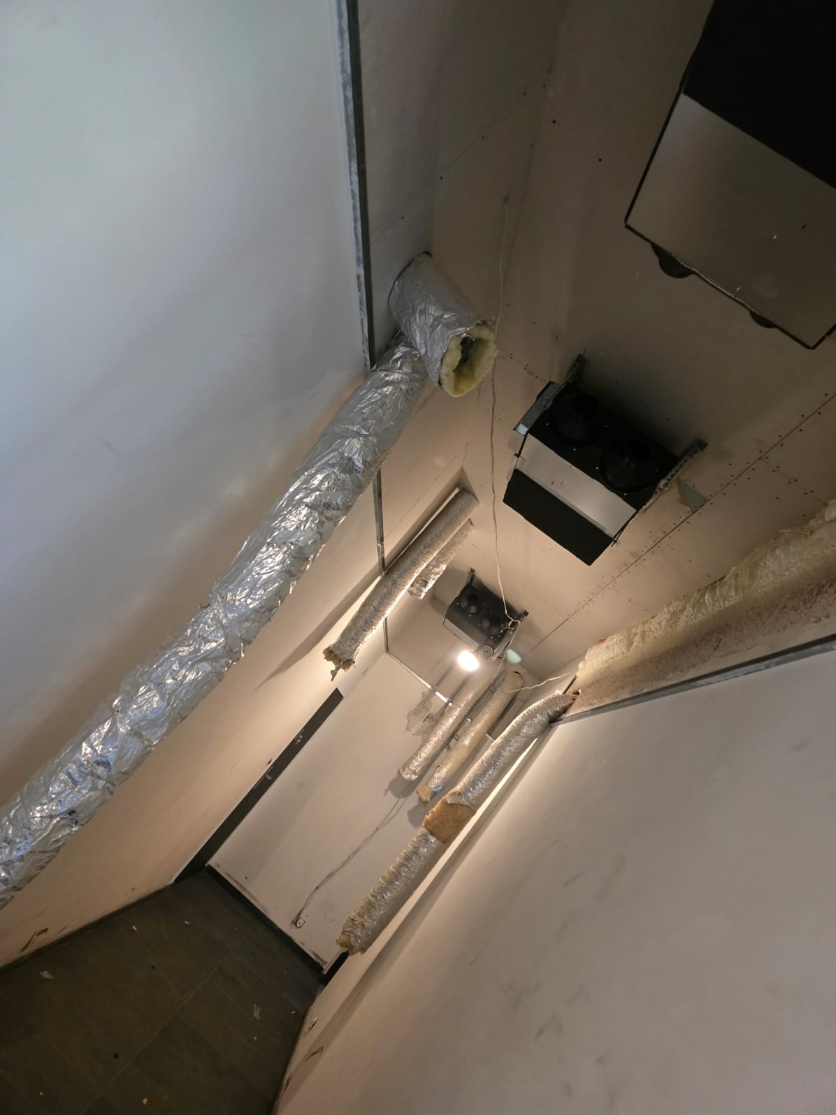 Instalacja wentylacyjna z aluminiowymi przewodami w trakcie montażu, widoczne elementy mocujące i izolacja, sufit z elementami oświetlenia i wentylacji, ściany w trakcie wykończenia.