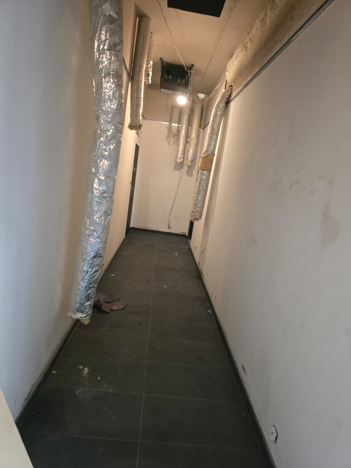 Korytarz w trakcie prac remontowych z widocznymi przewodami wentylacyjnymi owiniętymi folią, surowymi ścianami i ciemną podłogą z płytek.