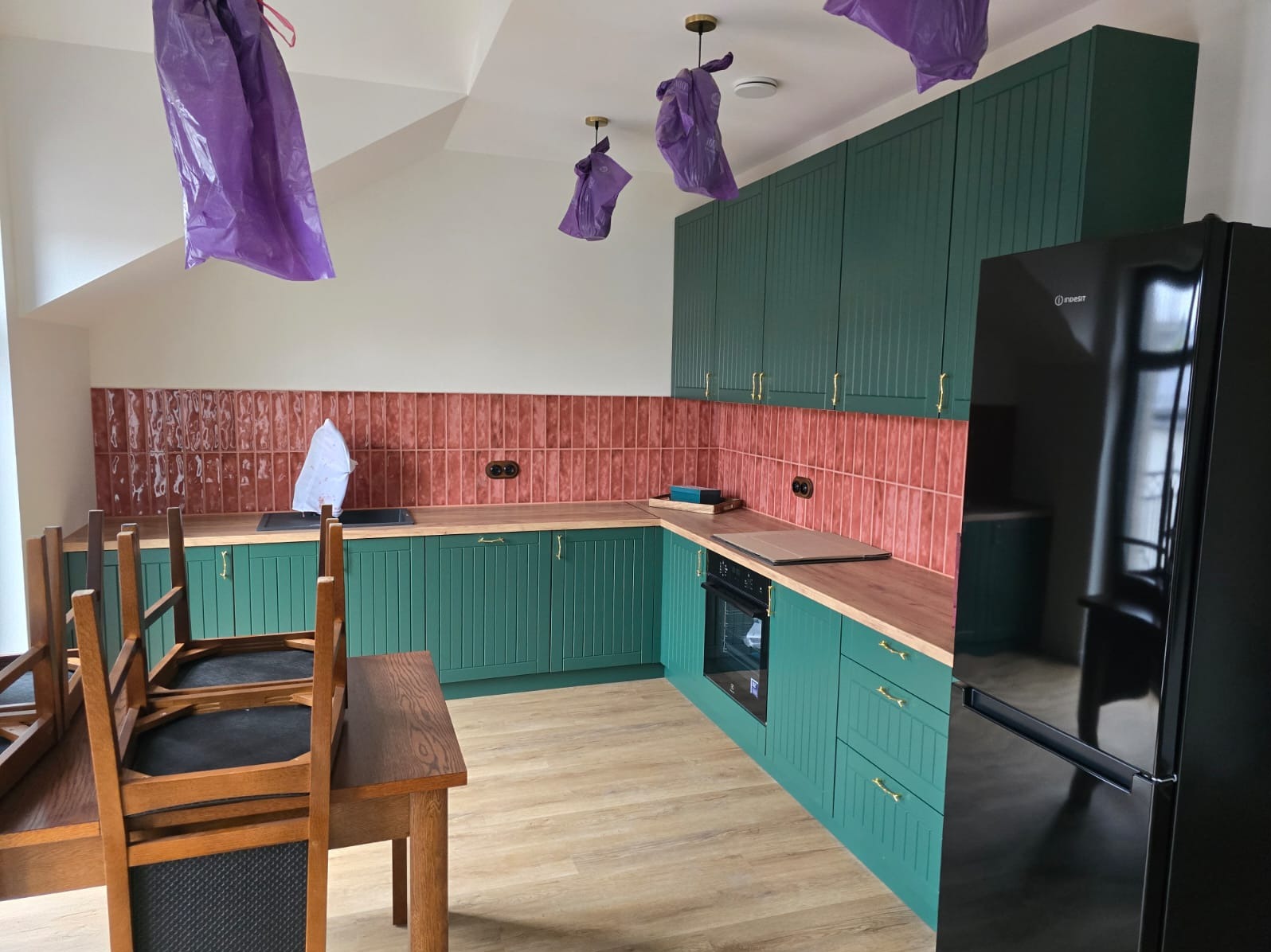 Nowoczesna kuchnia z zielonymi szafkami i ceglastymi płytkami, zabezpieczona folią podczas prac wykończeniowych, z widoczną czarną lodówką i drewnianym stołem z krzesłami.