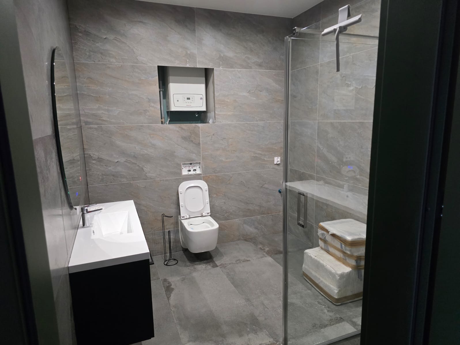 Nowoczesna łazienka z szarymi płytkami imitującymi kamień, wisząca toaleta z otwartą klapą, biała umywalka z czarną szafką, kabina prysznicowa ze szklaną ścianką i białe opakowania wewnątrz,...