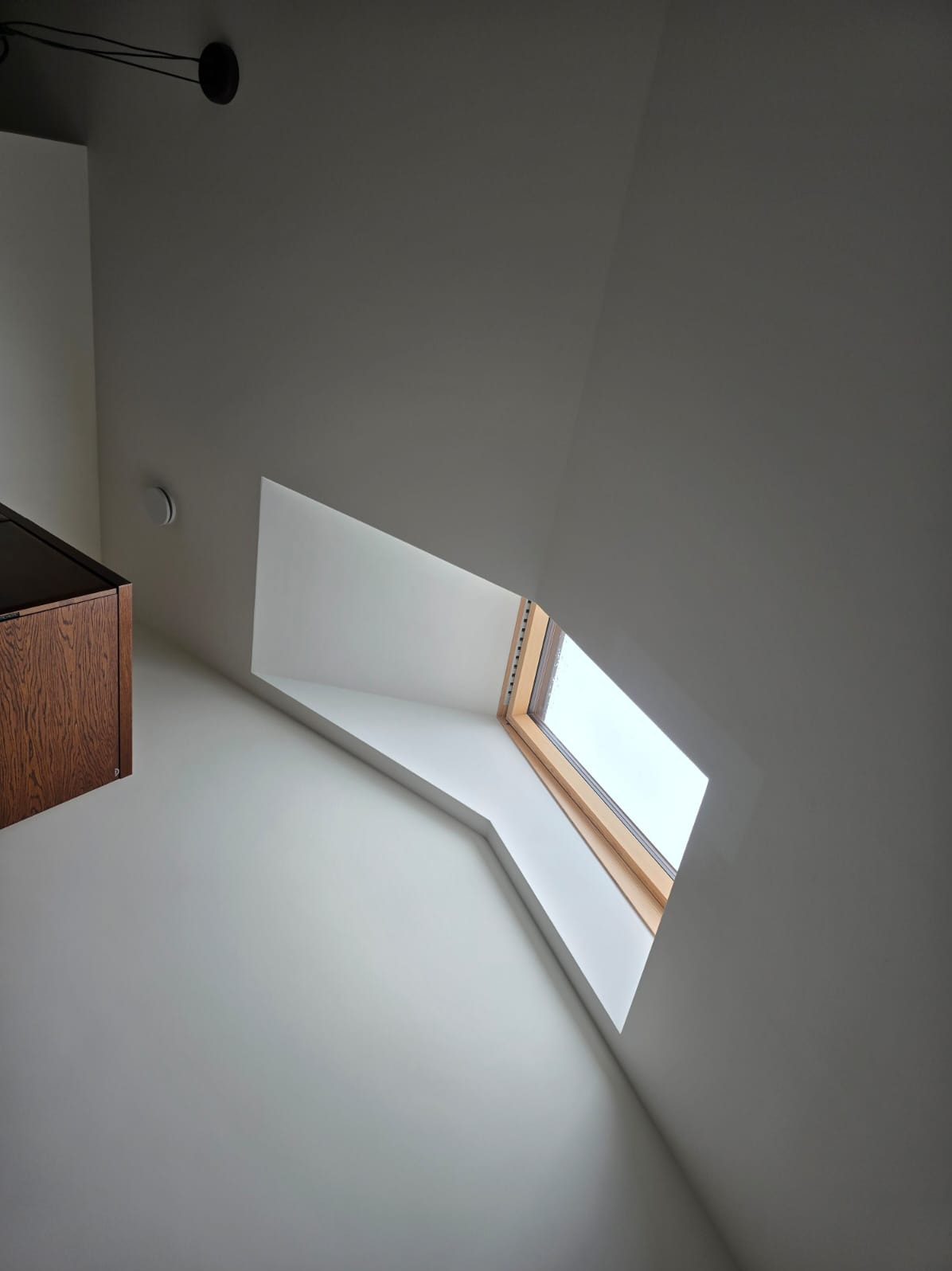 Poddasze z oknem dachowym w drewnianej ramie, białe ściany i fragment wiszącej szafki z ciemnego drewna, widok z dołu.