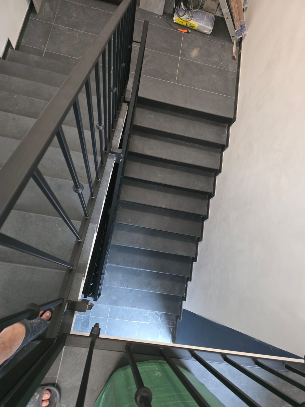 Schody wyłożone szarymi płytkami, z czarną metalową balustradą, widok z góry, częściowo widoczne stopy w klapkach, w tle wiadro i worki budowlane.