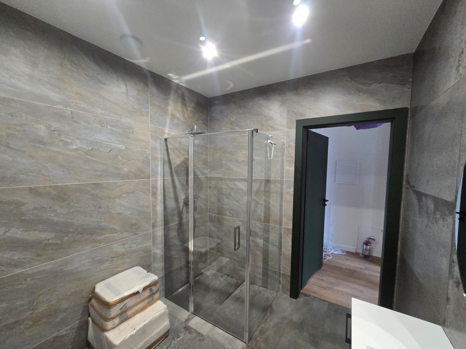 Nowoczesna kabina prysznicowa z szarego szkła, wykończona płytkami imitującymi kamień w odcieniach szarości i beżu, z widocznymi paczkami płytek czekającymi na montaż i otwartymi drzwiami...