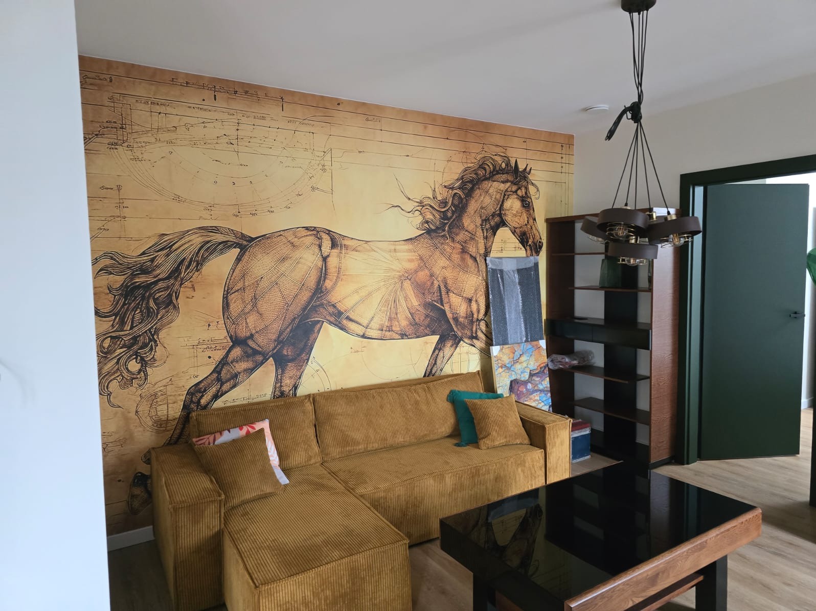Salon z sofą w kolorze musztardowym i fototapetą przedstawiającą konia w stylu szkiców Leonarda da Vinci, uzupełniony półkami i nowoczesną lampą wiszącą.