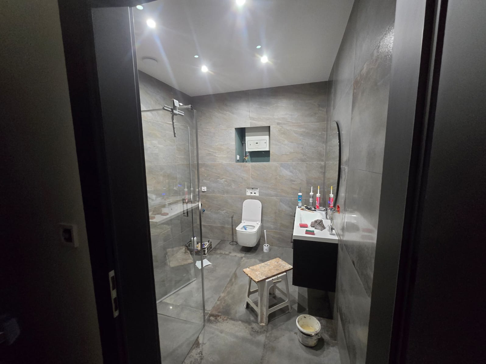 Nowoczesna łazienka z szarymi płytkami imitującymi kamień, kabiną prysznicową ze szkła, wiszącą toaletą i umywalką z szafką, widok na etap prac wykończeniowych z widocznym stołkiem i wiadrem.