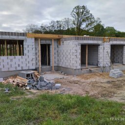 BUD Oleksandr Mishchur - Budowa domu jednorodzinnego z bloczków betonu komórkowego, widoczne otwory okienne i drzwiowe z drewnianymi belkami, teren budowy z materiałami i trawą.