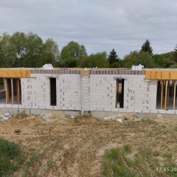 BUD Oleksandr Mishchur - Surowy stan budynku z betonu komórkowego w trakcie wznoszenia, widoczne otwory okienne i drzwiowe, drewniane szalunki i podpory, piaskowe podłoże z zielenią w tle.