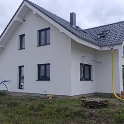 BUD Oleksandr Mishchur - Nowoczesny dom jednorodzinny z białą elewacją i czarną dachówką, widoczne przygotowania do podłączenia instalacji elektrycznej na ścianie, żółty wąż na elewacji, paleta i cegły na trawniku.