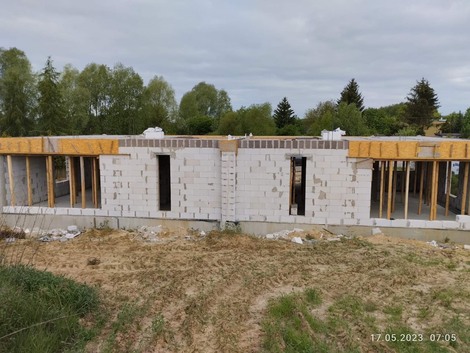 Surowy stan budynku z betonu komórkowego w trakcie wznoszenia, widoczne otwory okienne i drzwiowe, drewniane szalunki i podpory, piaskowe podłoże z zielenią w tle.