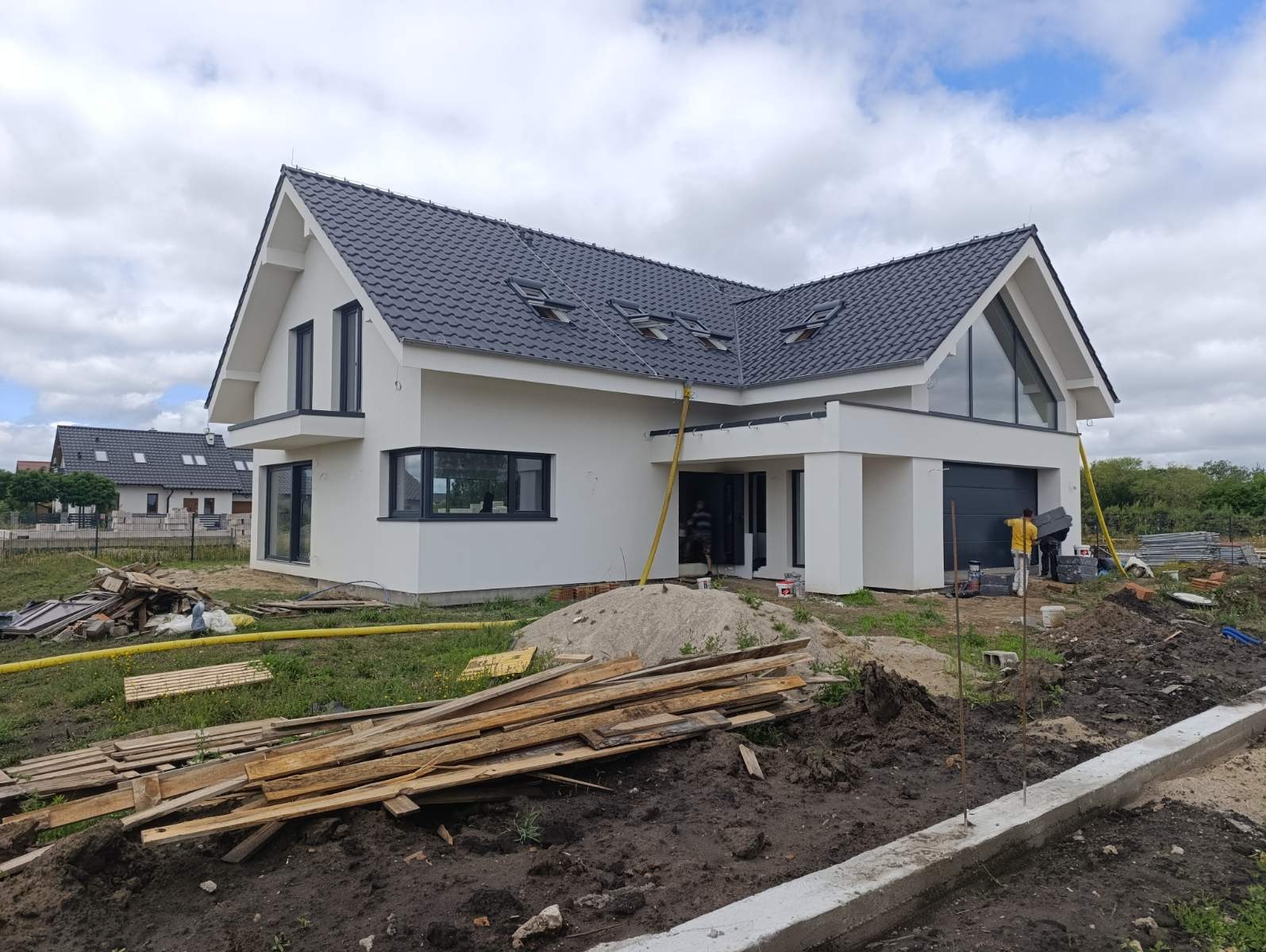 Nowoczesny dom jednorodzinny z białą elewacją i grafitowym dachem z lukarnami, widoczne rusztowania i materiały budowlane na placu budowy, pracownicy w trakcie prac wykończeniowych.