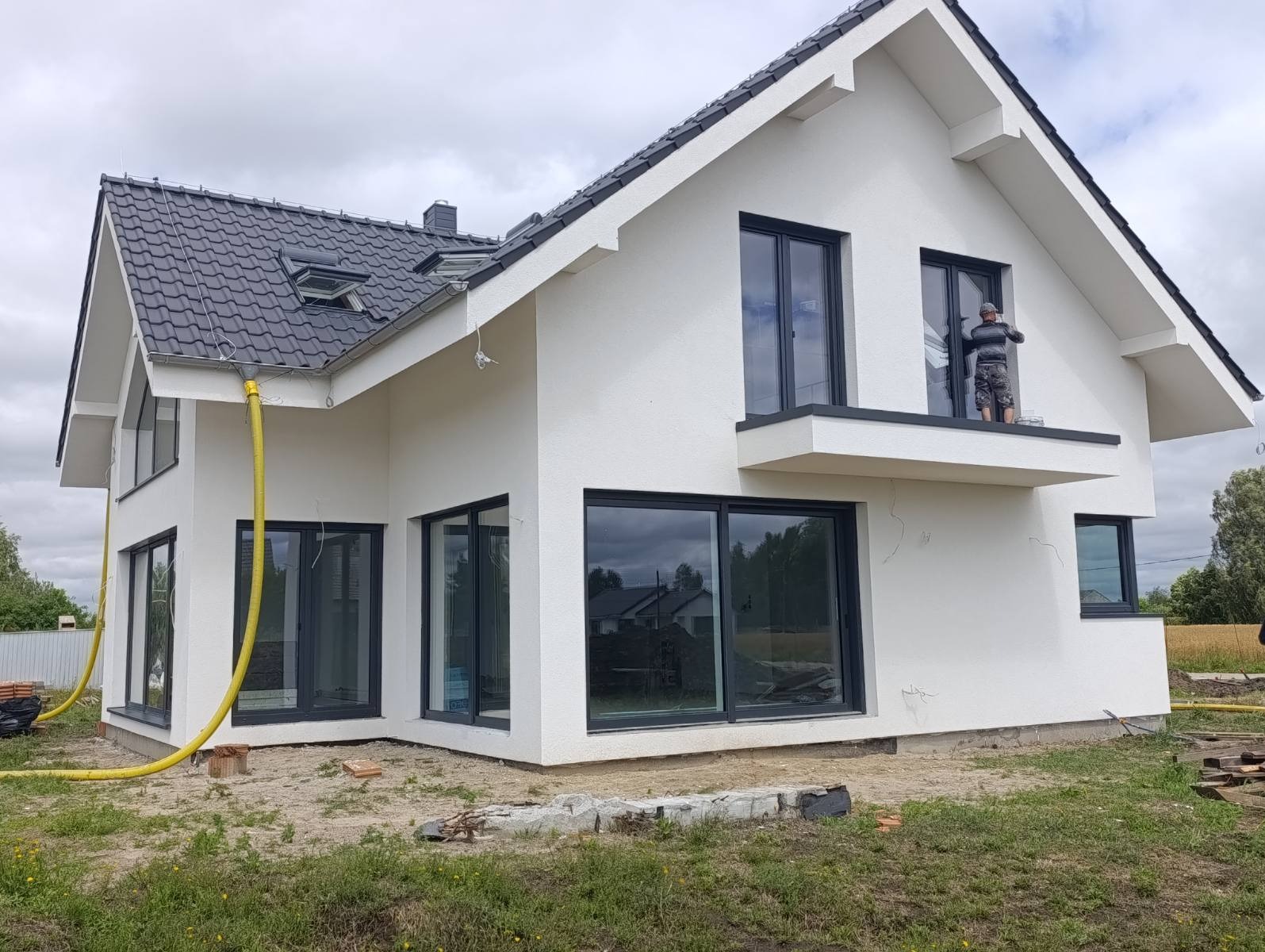 Nowoczesny dom jednorodzinny po tynkowaniu elewacji, z widocznym pracownikiem na balkonie i żółtym wężem do tynkowania leżącym na ziemi.