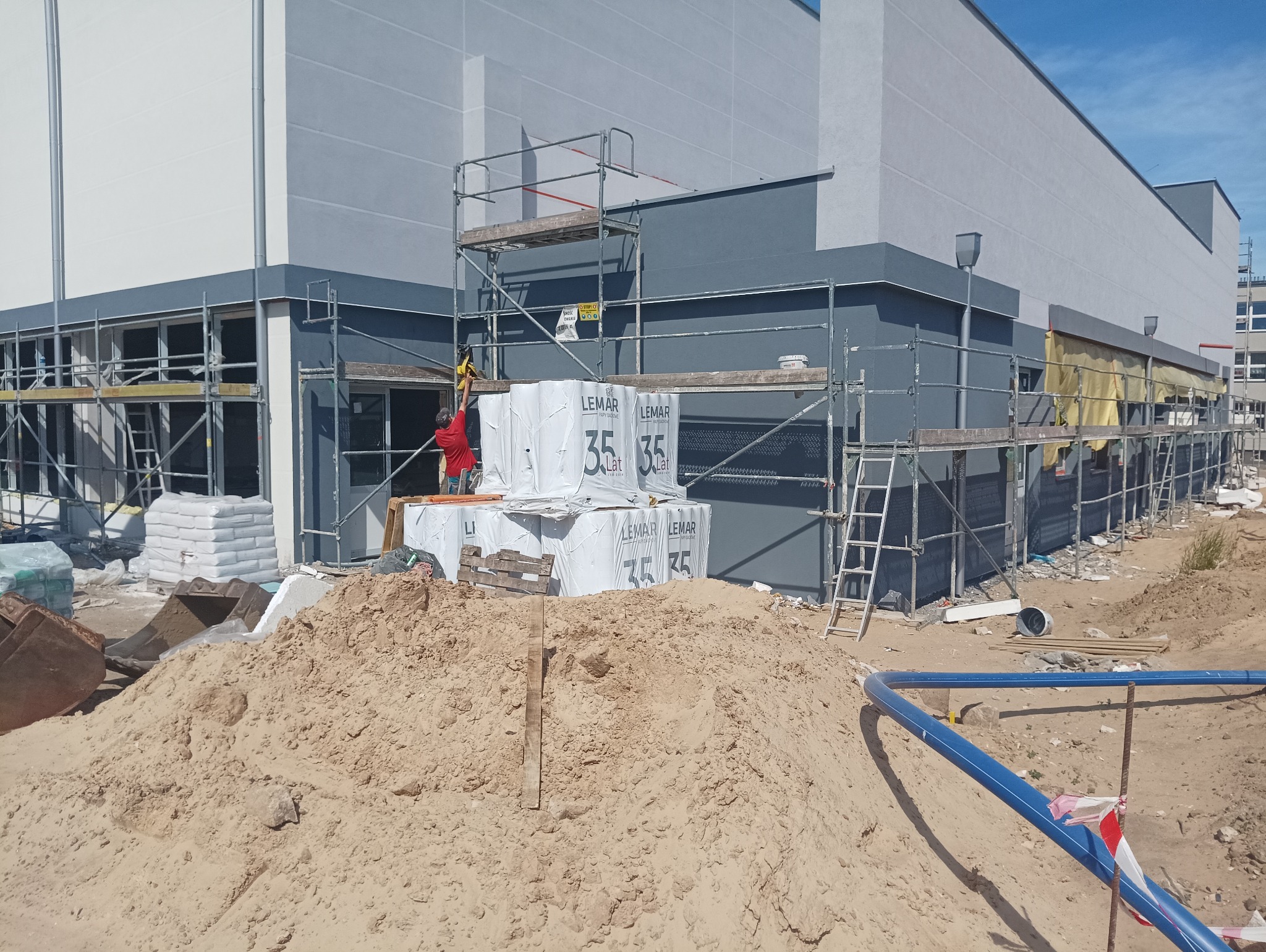 Budynek w trakcie prac elewacyjnych: widoczne rusztowania, sterty materiałów budowlanych marki Lemar, oraz pracownik w czerwonej koszulce pracujący na rusztowaniu.