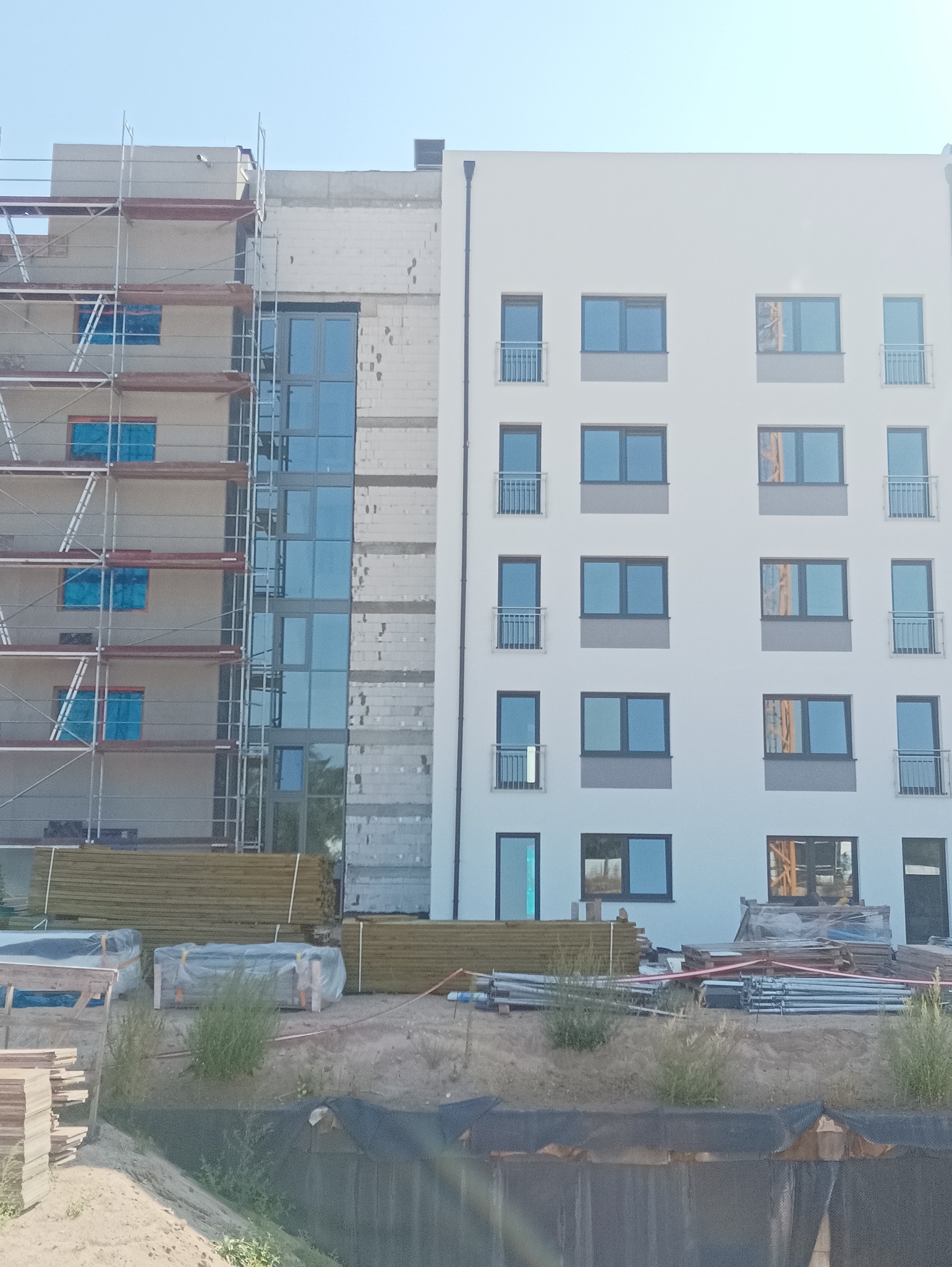 Budynek wielorodzinny w trakcie ocieplania elewacji, widoczne rusztowania, część elewacji otynkowana i pomalowana na biało, okna zabezpieczone folią, materiały budowlane składowane na placu budowy.