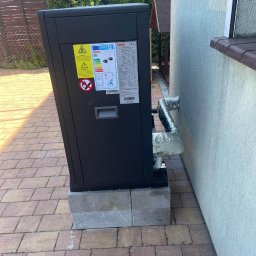 UNIQUE Karol Rupotacki - Czarna pompa ciepła postawiona na betonowych bloczkach przy ścianie budynku, z widocznymi rurami w izolacji, na tle kostki brukowej i ogrodzenia.