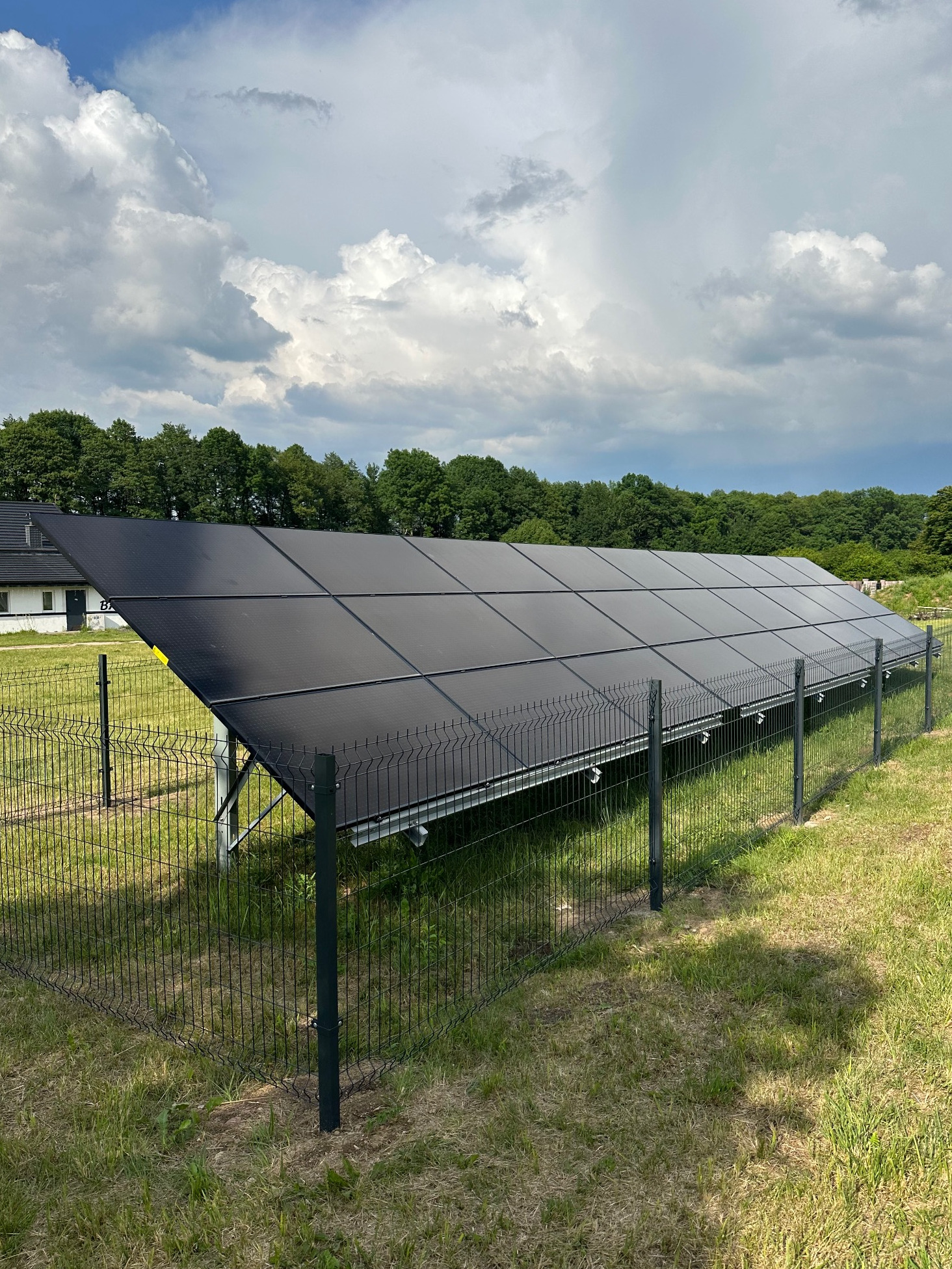 Rząd czarnych paneli słonecznych za ogrodzeniem z siatki na zielonym trawniku, z budynkiem i drzewami w tle pod pochmurnym niebem.