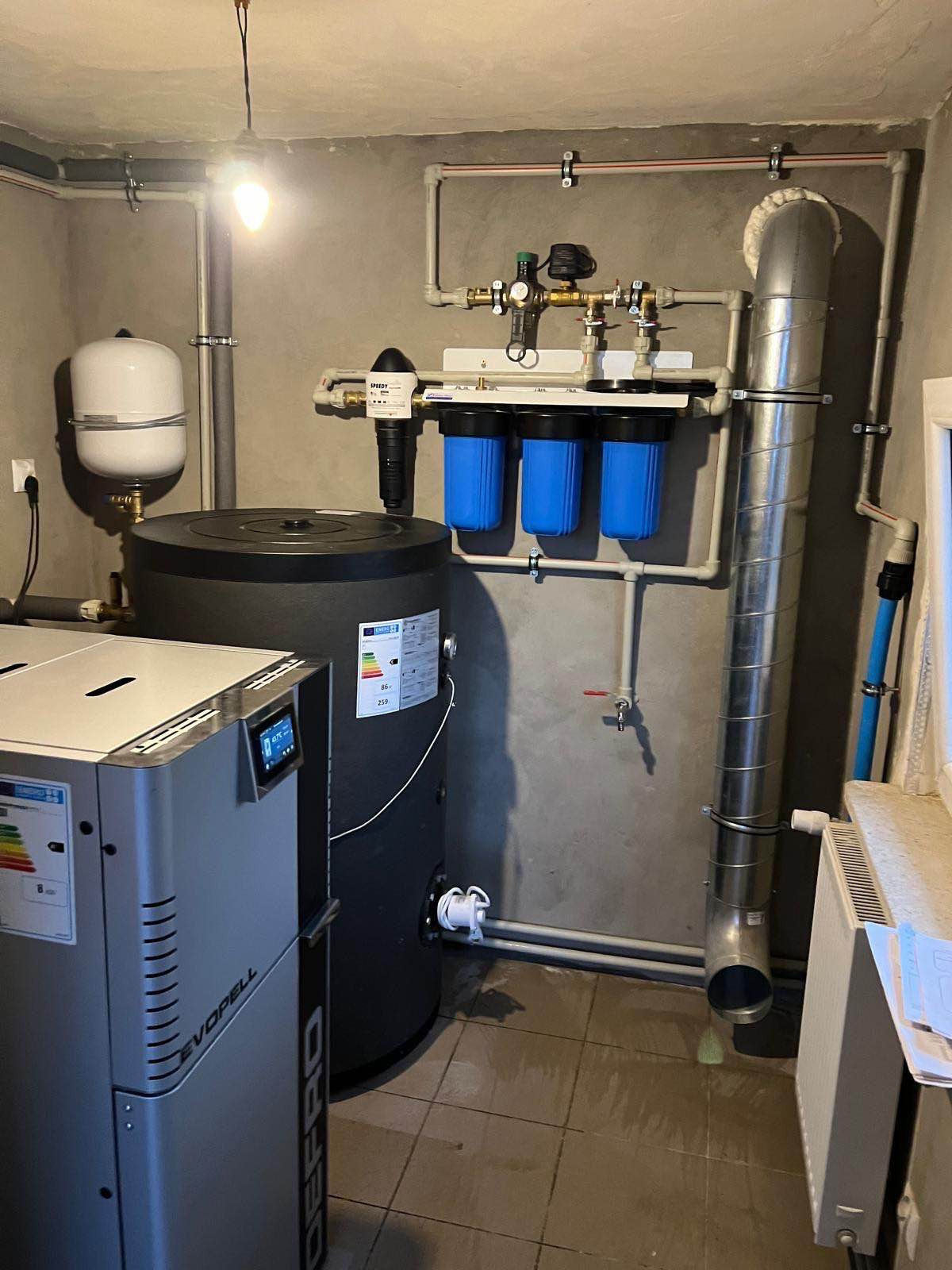 Kotłownia z kotłem na pellet Defro Evopell, zbiornikiem buforowym, filtrami wody z niebieskimi wkładami i instalacją grzewczą z rurami PCV.
