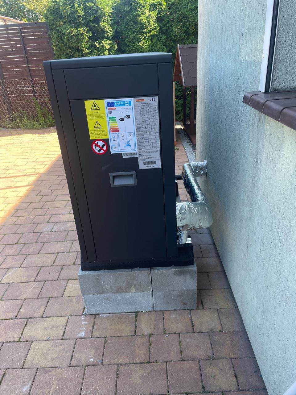 Czarna pompa ciepła postawiona na betonowych bloczkach przy ścianie budynku, z widocznymi rurami w izolacji, na tle kostki brukowej i ogrodzenia.