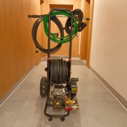 Przenośne urządzenie WUKO z nawiniętym wężem ciśnieniowym, gotowe do udrażniania rur wewnątrz budynku. Widoczne złącza i dysze. Szary korytarz w tle.