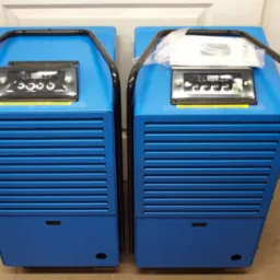 Dwa niebieskie osuszacze powietrza z czarnymi uchwytami i panelem sterowania, naklejka z numerem telefonu. Urządzenia gotowe do użycia.