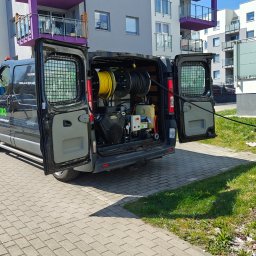 Serwis Techniczny Trójmiasto - Czarny van z otwartymi drzwiami, wewnątrz sprzęt do czyszczenia kanalizacji. Wąż prowadzi do studzienki na trawniku przed blokiem mieszkalnym.