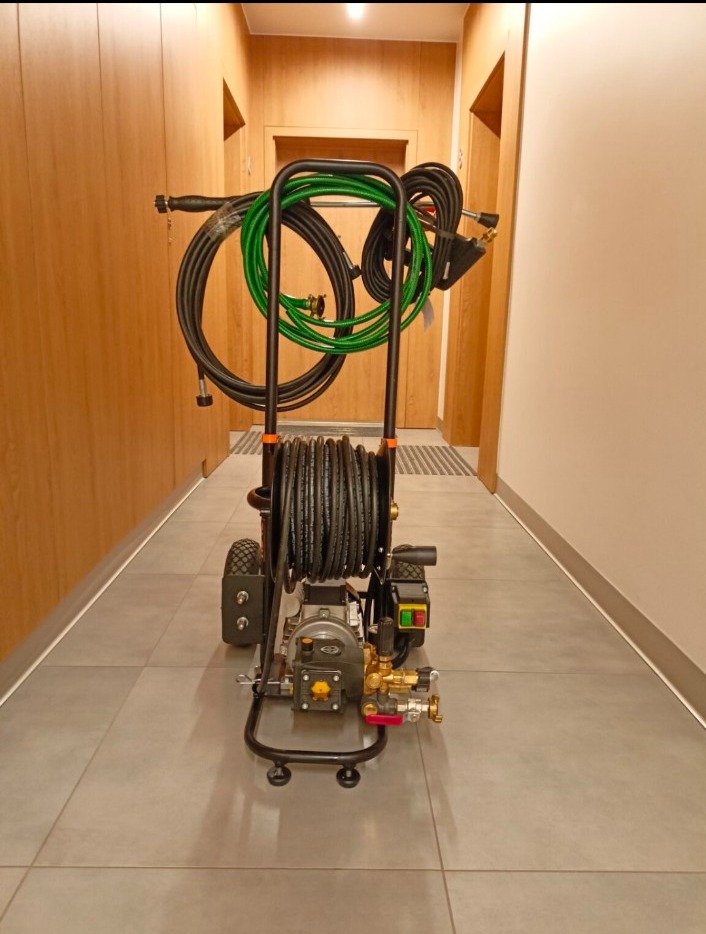 Przenośne urządzenie WUKO z nawiniętym wężem ciśnieniowym, gotowe do udrażniania rur wewnątrz budynku. Widoczne złącza i dysze. Szary korytarz w tle.