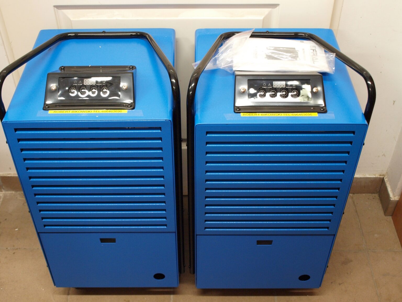 Dwa niebieskie osuszacze powietrza z czarnymi uchwytami i panelem sterowania, naklejka z numerem telefonu. Urządzenia gotowe do użycia.