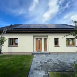 Jednorodzinny dom z beżową elewacją, brązowymi oknami i drzwiami oraz zamontowanymi panelami słonecznymi na dachu. Aluminiowa drabina oparta o dach. Brukowany podjazd.