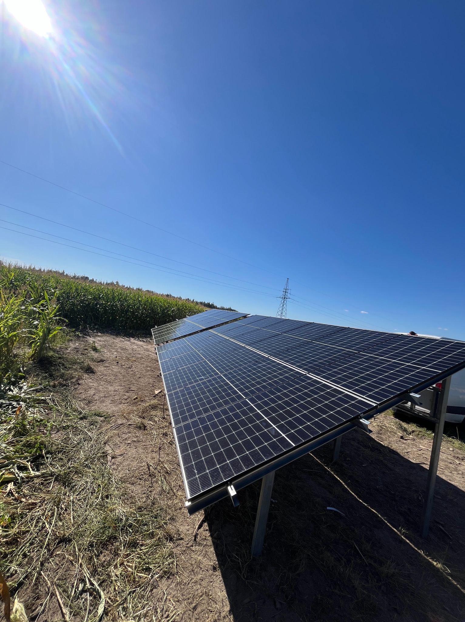 Rząd paneli słonecznych ustawionych na polu, z widoczną linią wysokiego napięcia i polem kukurydzy w tle, pod błękitnym niebem z promieniami słońca.