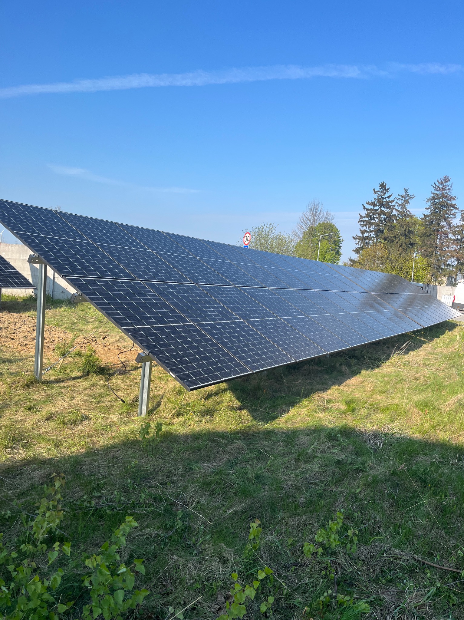 Rząd paneli słonecznych na metalowych stelażach, zamontowanych na trawiastym terenie w słoneczny dzień, widoczny znak drogowy w tle.