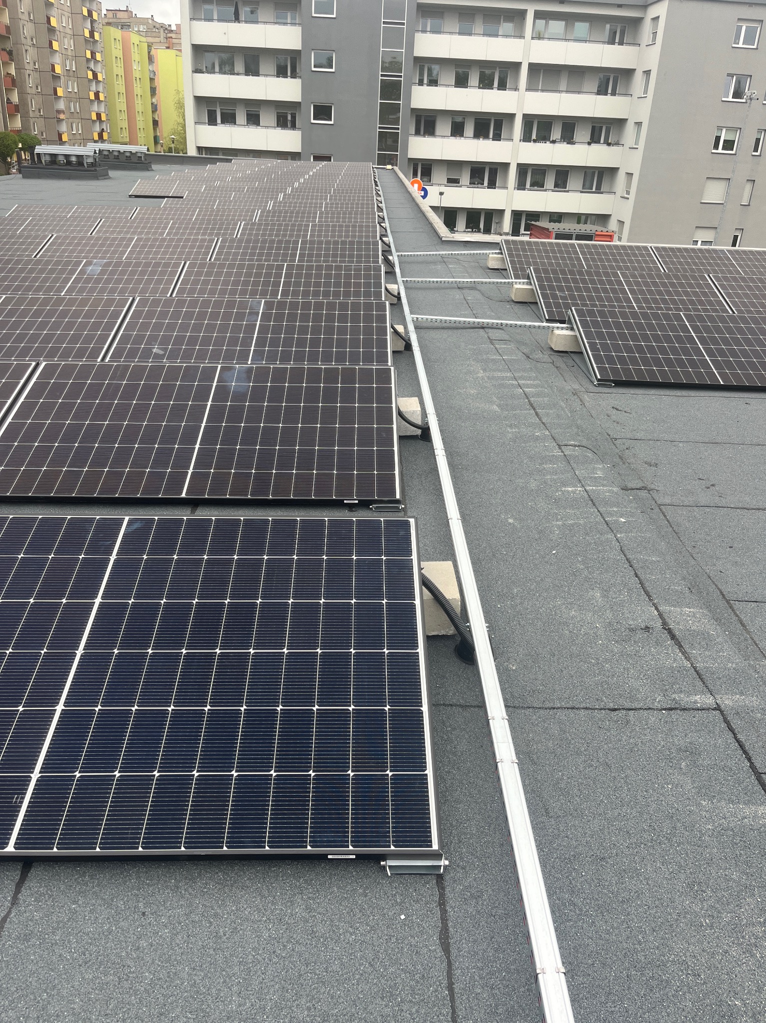 Rząd paneli słonecznych z widoczną konstrukcją montażową na płaskim, bitumicznym dachu budynku wielorodzinnego w pochmurny dzień.