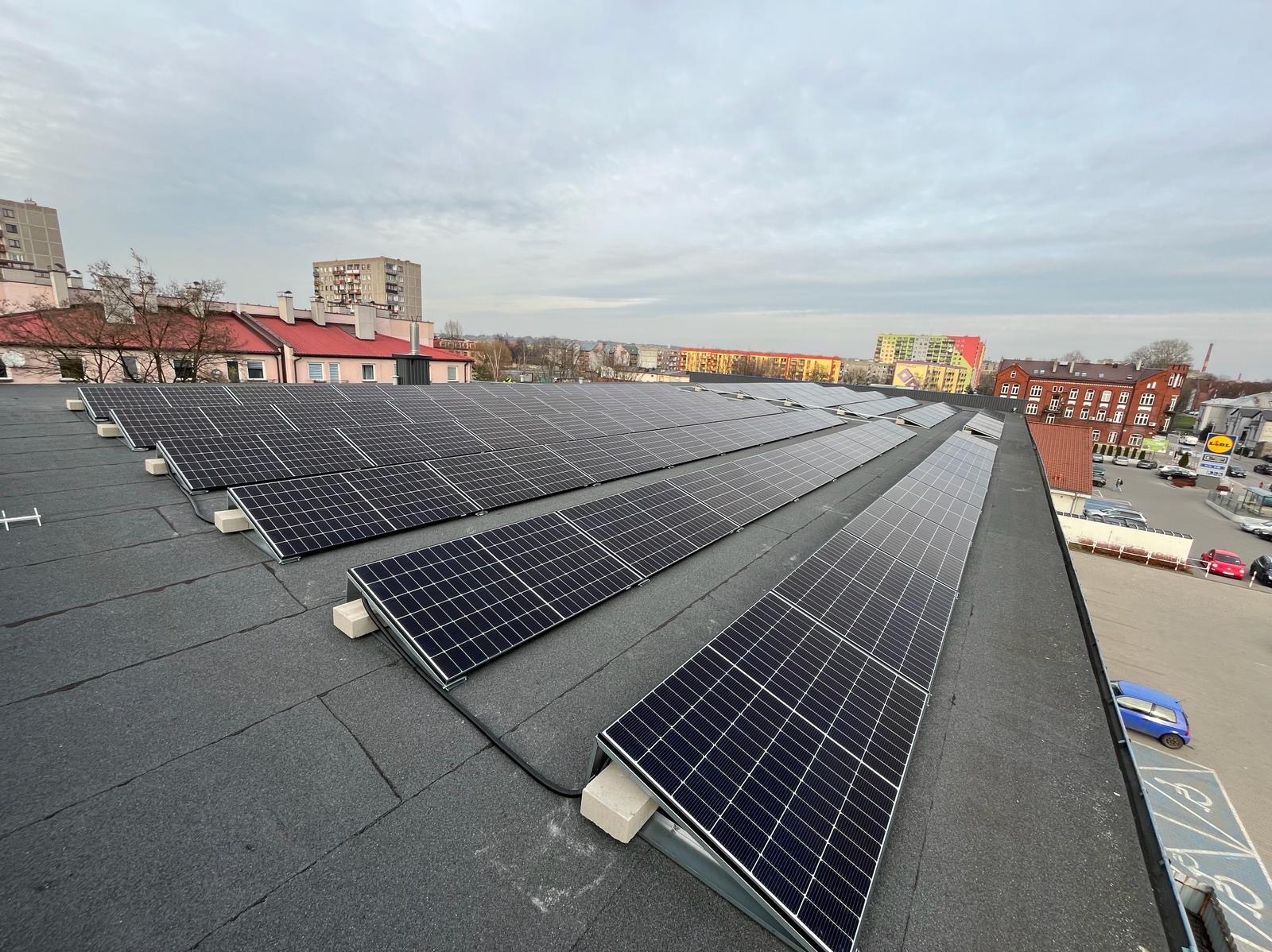 Rzędy paneli słonecznych na płaskim dachu pokrytym papą, z widokiem na miejski krajobraz w pochmurny dzień.