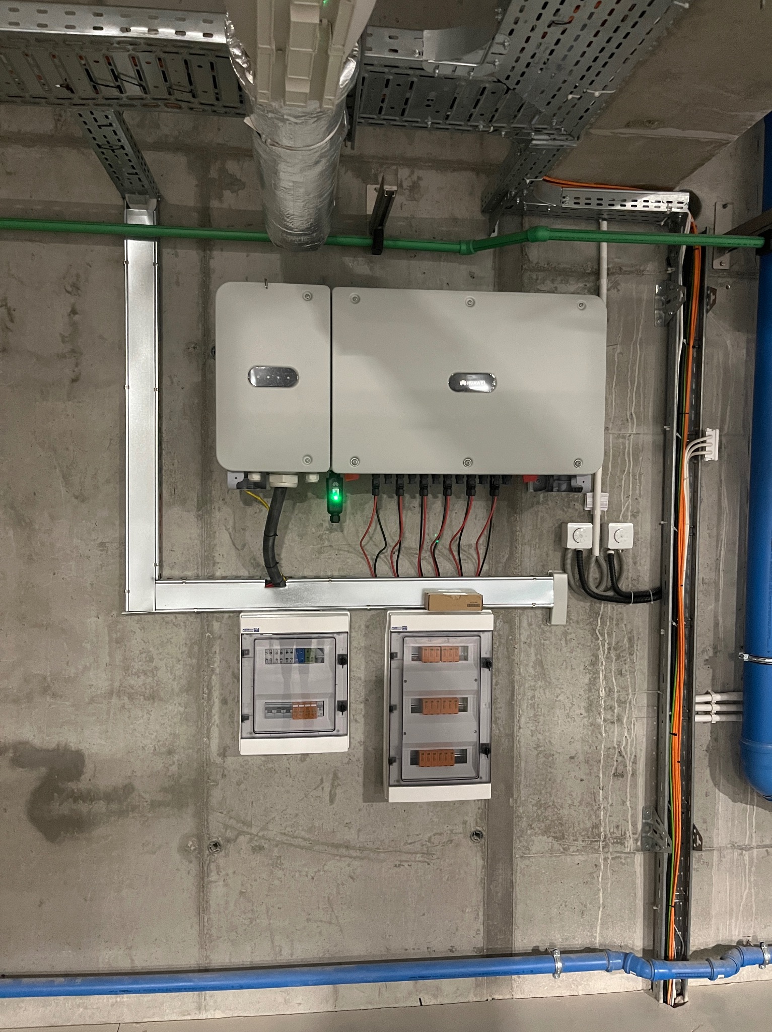 Dwa inwertery fotowoltaiczne marki Huawei zamontowane na szarej, betonowej ścianie, poniżej dwie skrzynki z bezpiecznikami, widoczne kable i rury instalacyjne.