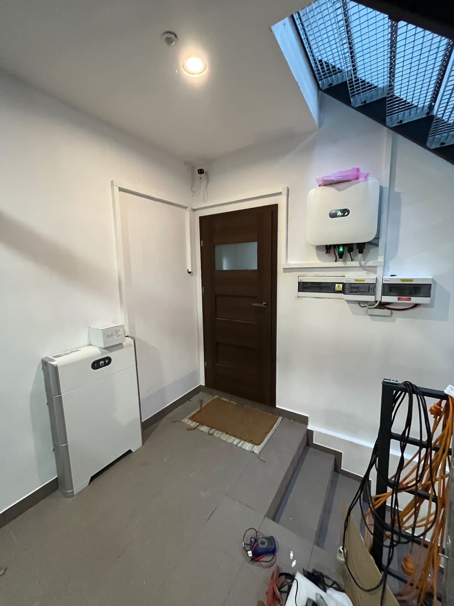 Wnętrze pomieszczenia z zainstalowanym magazynem energii, inwerterem fotowoltaicznym i rozdzielnicą elektryczną przy schodach. Na podłodze leżą narzędzia pomiarowe.