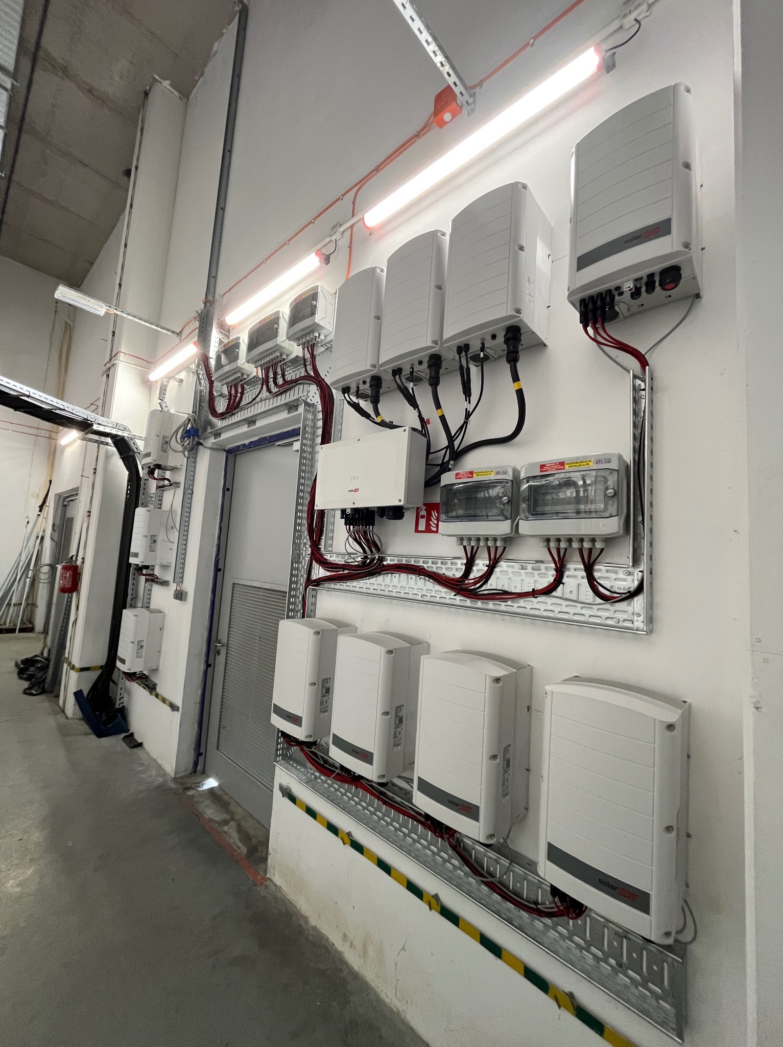 Rząd białych inwerterów zamontowanych na ścianie w pomieszczeniu technicznym, z widocznymi przewodami i elementami instalacji elektrycznej.