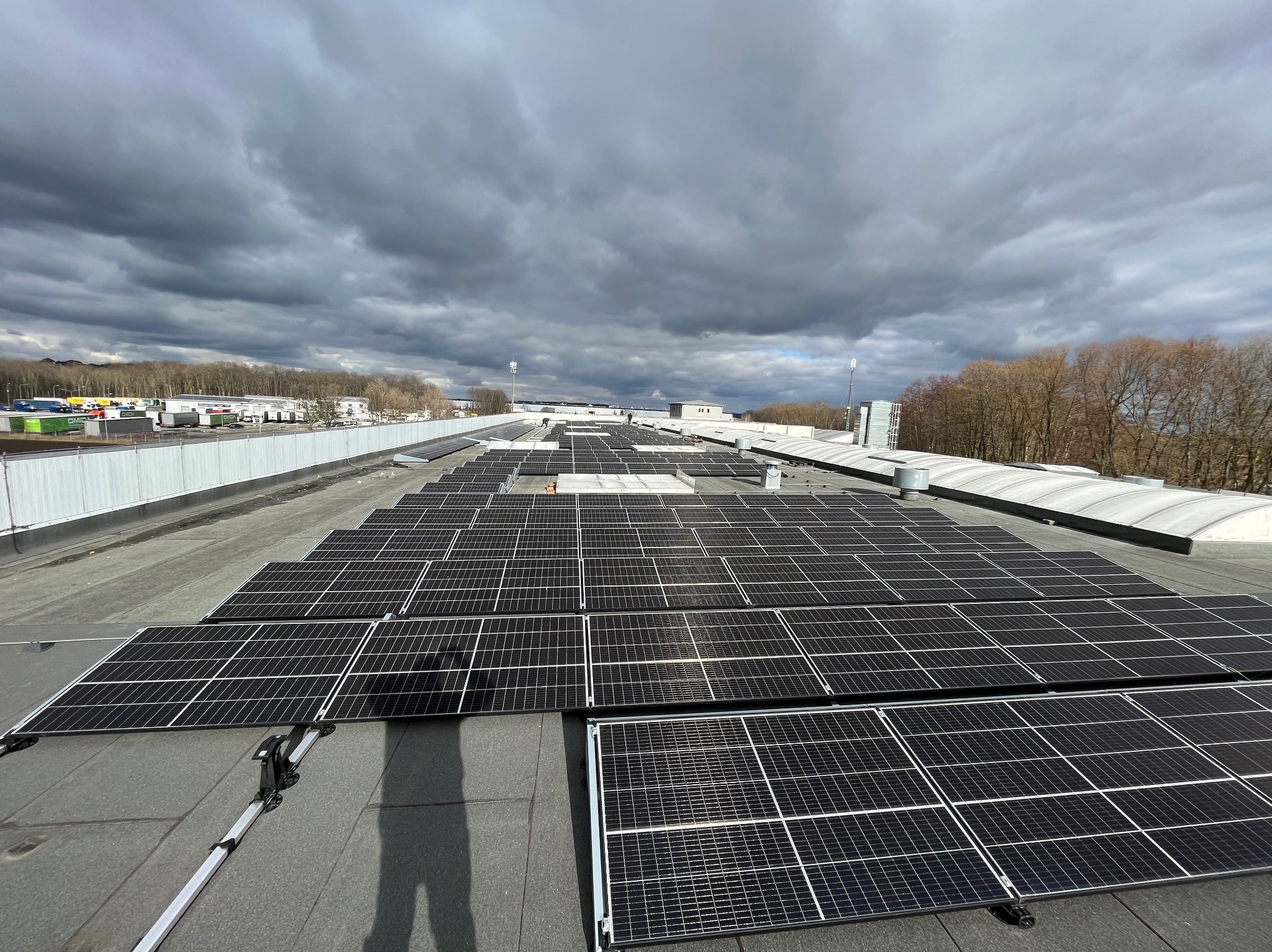 Rozległa instalacja paneli słonecznych na płaskim dachu budynku przemysłowego, widok z góry, pochmurne niebo w tle.