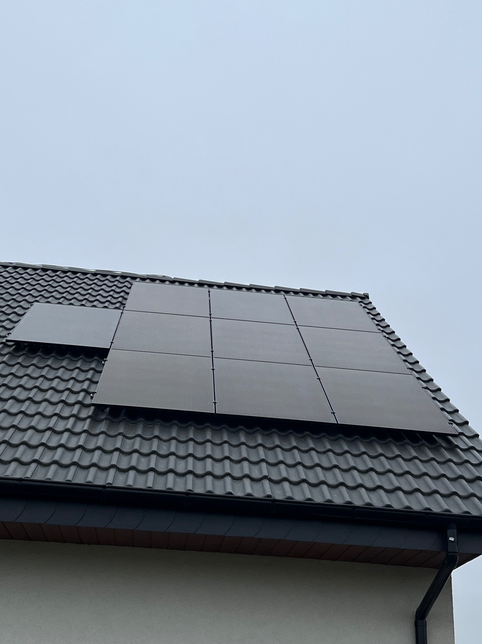 Częściowy widok paneli słonecznych na dachu z szarej dachówki ceramicznej, pochmurne niebo w tle.