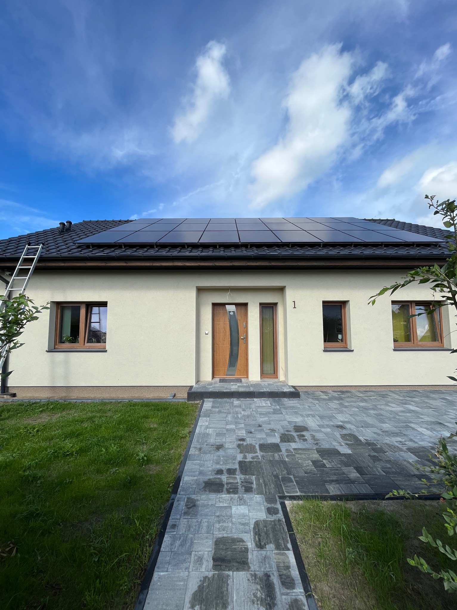 Jednorodzinny dom z beżową elewacją, brązowymi oknami i drzwiami oraz zamontowanymi panelami słonecznymi na dachu. Aluminiowa drabina oparta o dach. Brukowany podjazd.