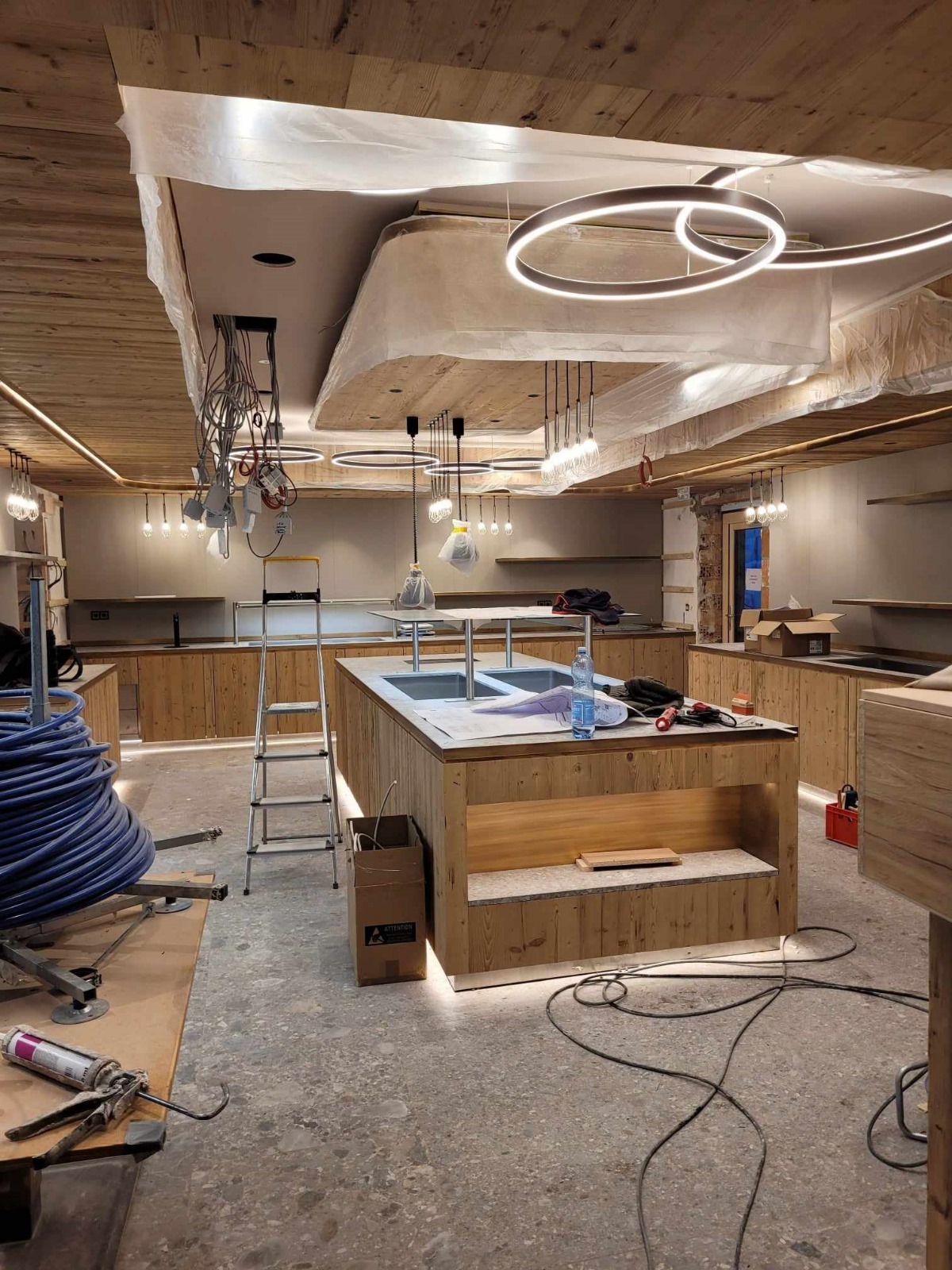 Wnętrze lokalu w trakcie wykańczania z drewnianą zabudową, wyspą z dwoma zlewozmywakami i wiszącymi lampami LED, kable zwisające z sufitu, drabina i narzędzia na podłodze.