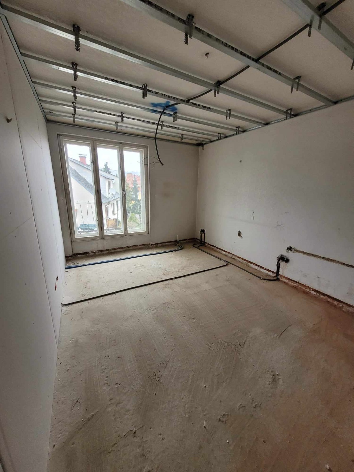 Wnętrze w trakcie remontu: widoczne profile sufitowe, okno z widokiem na dom, kable wystające ze ściany i sufitu oraz częściowo wykończone ściany z płyt gipsowo-kartonowych.