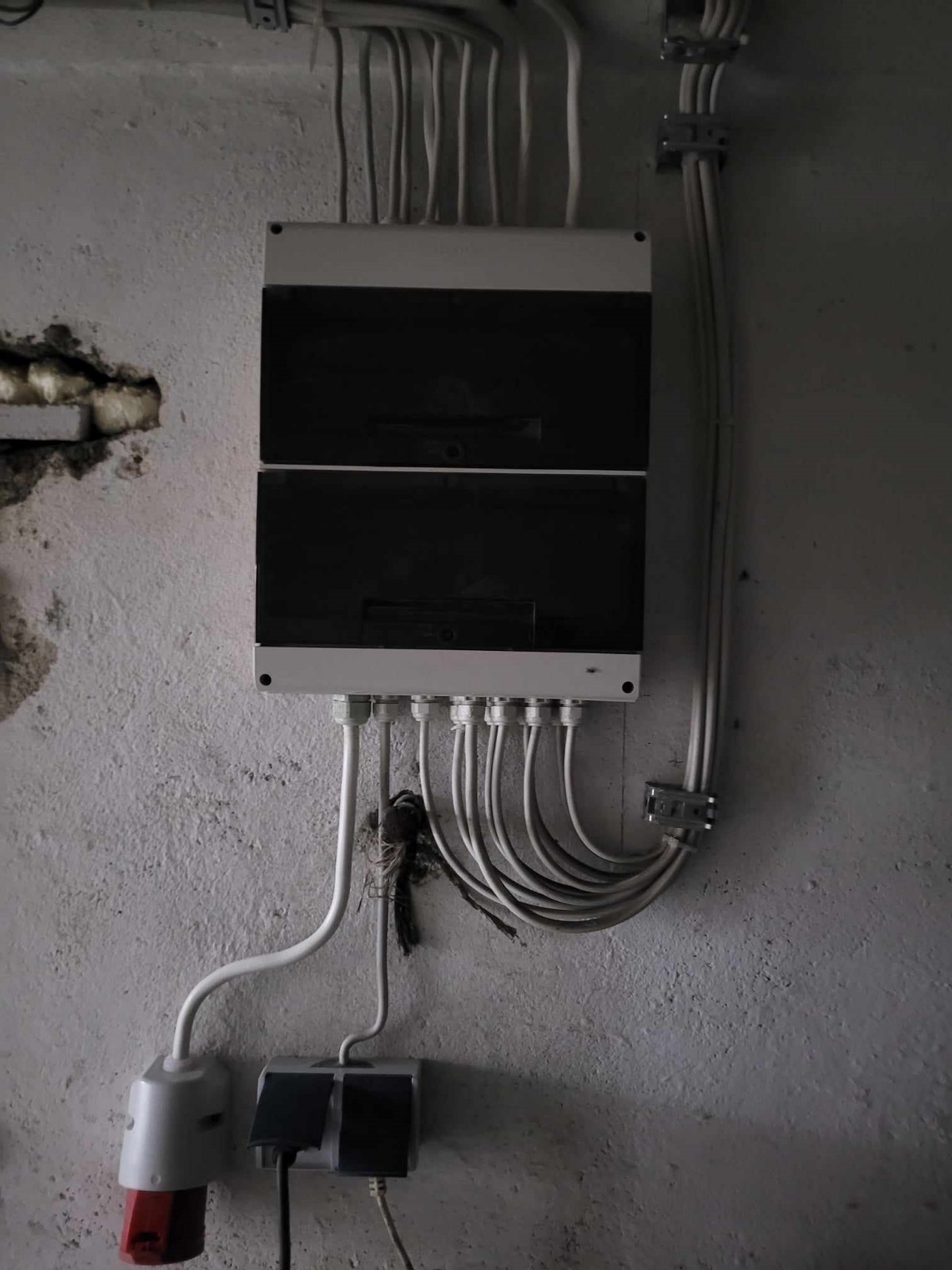 Szara, betonowa ściana z zamontowaną, dwuczęściową, biało-czarną skrzynką elektryczną, z której wychodzą liczne kable w białych osłonach, dodatkowo widoczny element przyłącza z czerwonym akcentem.