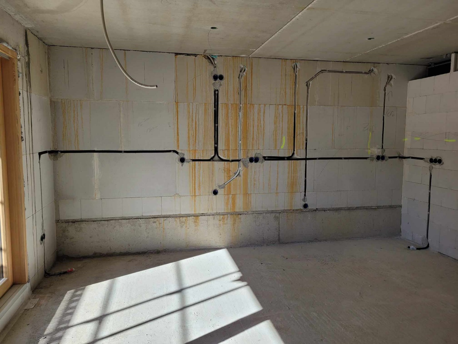 Surowy pokój w trakcie instalacji elektrycznej: czarne kable prowadzone po ścianach z betonu komórkowego, widoczne puszki instalacyjne, ślady po zaciekach na ścianach, wpadające światło słoneczne.