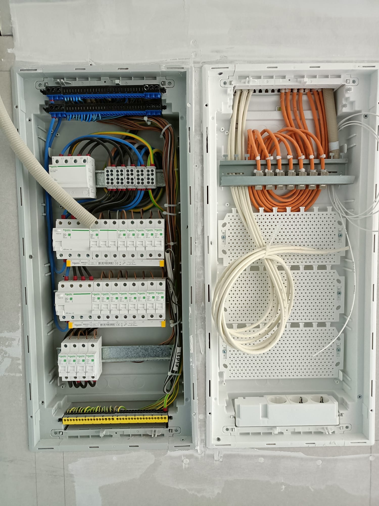 Otwarta skrzynka rozdzielcza z zabezpieczeniami, kablami w kolorach niebieskim, żółtym, pomarańczowym i brązowym, zamontowana w ścianie w trakcie wykończenia.