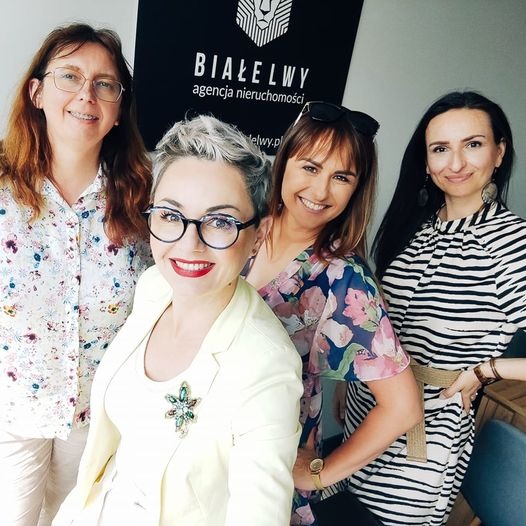 Portret czterech uśmiechniętych kobiet, pracowniczek agencji nieruchomości 'Białelwy', na tle ściany z logo firmy.