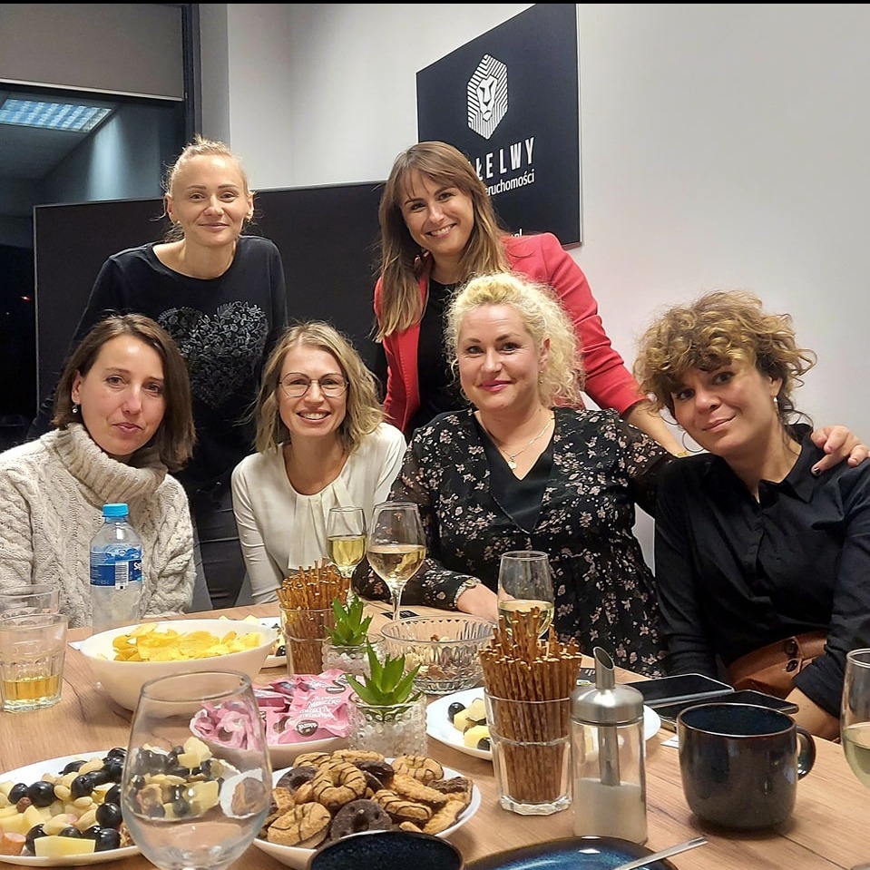 Grupa sześciu uśmiechniętych kobiet siedzi przy stole zastawionym przekąskami, winem i słodyczami, w tle widoczne logo agencji nieruchomości.